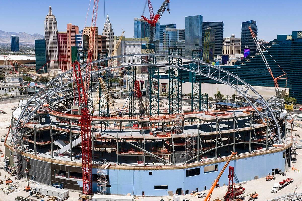 Las Vegas stadium construction with curved steel roof framing, cranes and the Strip skyline behind it